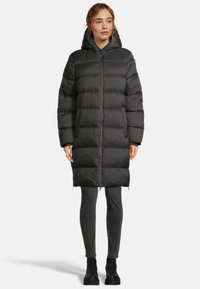 Femme portant un long manteau rembourré noir avec capuche, un pantalon skinny noir et des bottes à plateforme noires, se tenant devant un fond uni.