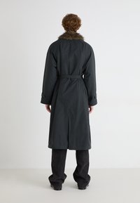 Trench coat gri închis cu guler de blană, talie cu cordon și manșete cu nasturi. Purtați deasupra unor pantaloni negri cu picior larg, văzuți din spate.