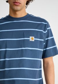 Námořnická modrá tričko s krátkým rukávem se světle modrými horizontálními pruhy, kapsou na prsou a štítkem s logem Carhartt na kapse.