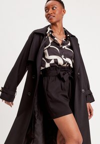 Sort trenchcoat over en mønstret bluse i sort og creme, parret med højtaljede sorte shorts med bælte i taljen.