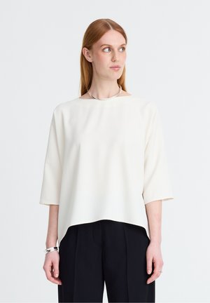 Femme aux cheveux roux lisses portant un haut blanc ample à manches trois-quarts et un pantalon noir, regardant vers sa droite.