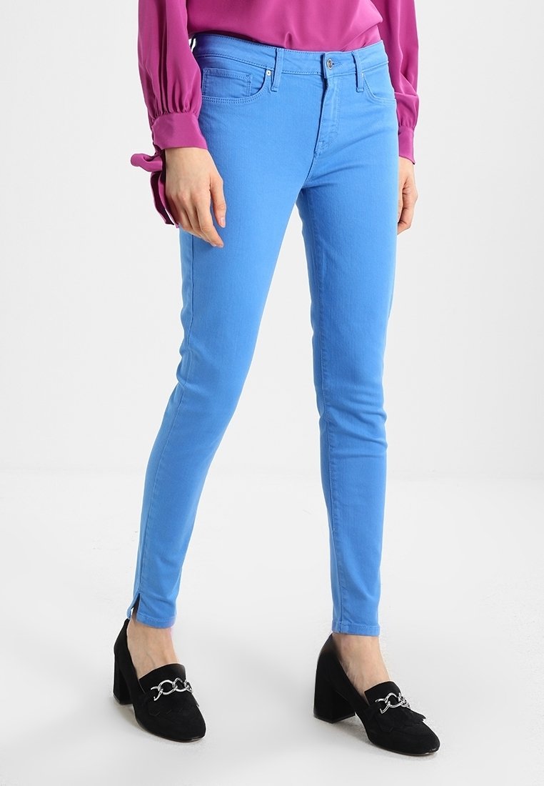 Vaqueros ajustados de un brillante color azul con una textura suave, que presentan un diseño estándar de cinco bolsillos y aberturas en los tobillos, combinados con zapatos de tacón negros.