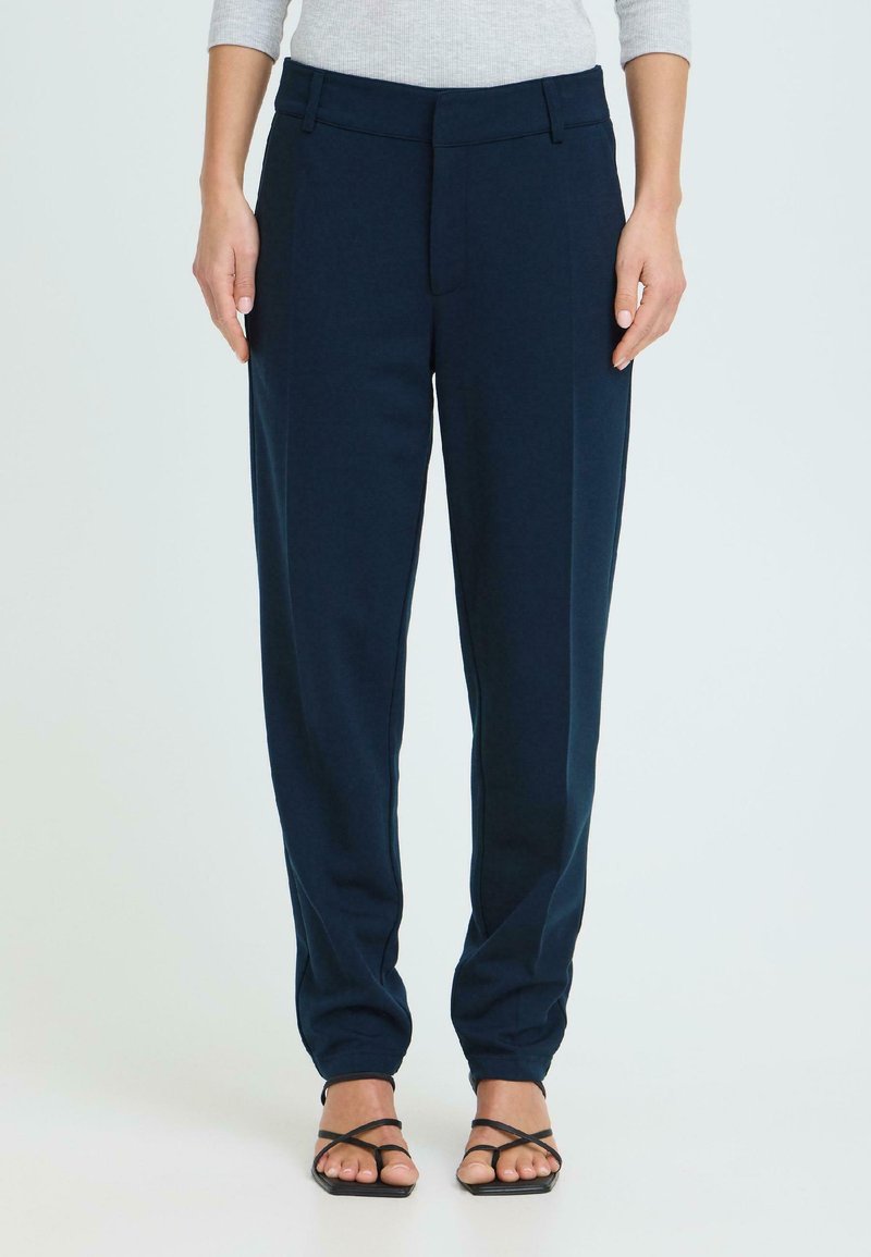Femme portant un pantalon droit bleu marine avec un pli à l'avant, associé à un haut à manches longues gris clair et des sandales noires à lanières.