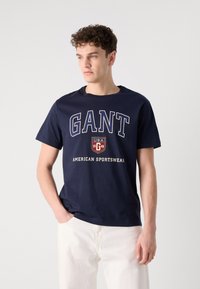 Tricou din bumbac de culoare bleumarin, cu un logo alb îndrăzneț "GANT", un design cu scut și textul "AMERICAN SPORTSWEAR". Guler clasic tip crew și mâneci scurte.