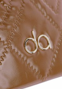 Superficie de cuero marrón brillante con costuras decorativas y un logo metálico dorado. La bolsa tiene una textura suave con un diseño moderno.