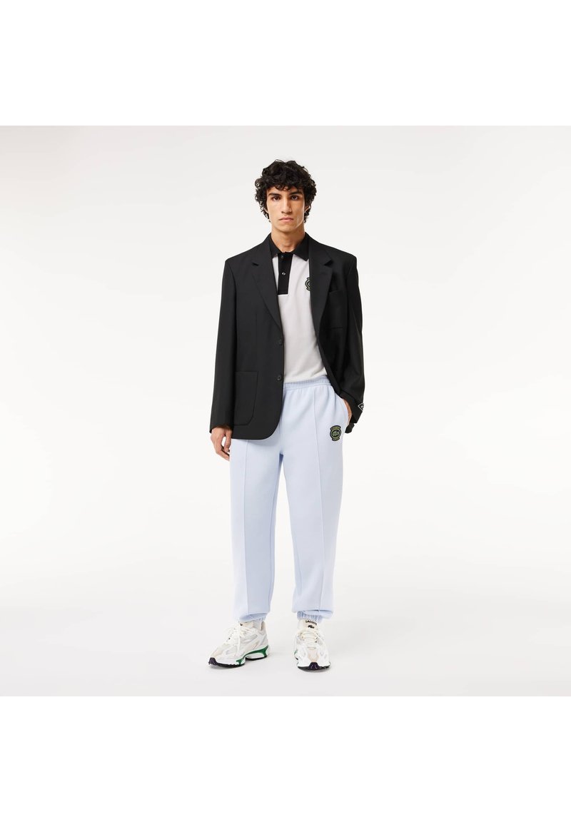 Blazer noir porté sur un polo blanc et noir, jogging bleu clair avec un logo, et chaussures blanches avec des accents verts et noirs.