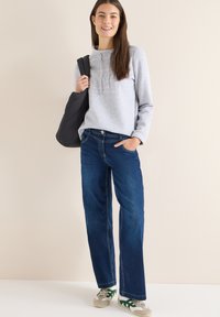 Felpa con cappuccio grigio chiaro con coulisse, jeans blu a gamba larga e sneakers grigie con dettagli verdi. Modella che tiene una borsa a tracolla nera.