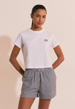 Mujer con el pelo trenzado que lleva una camiseta blanca de manga corta con un pequeño logo y pantalones cortos de cuadros en blanco y negro de talle alto, con las manos en los bolsillos.