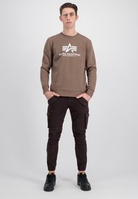 Sudadera marrón con logo blanco, combinada con pantalones cargo marrones oscuros con bolsillos y puños elásticos. Zapatillas negras completan el atuendo.