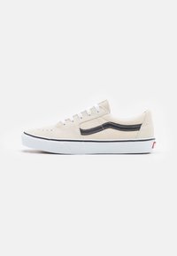 Beige suede sneaker with a black side stripe, white laces, and rubber sole. Features perforations on the toe and a low-profile design.