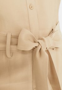 Haut beige ceinturé présentant un tissu texturé, une fermeture à bouton et un détail de nœud sur le devant pour un ajustement réglable.