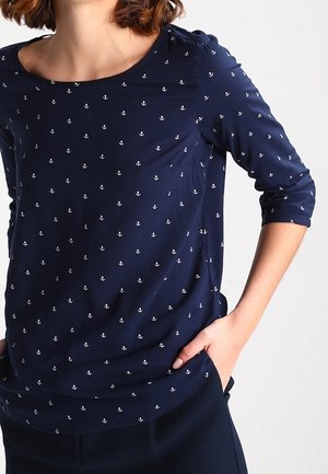 Kvinde iført en marineblå langærmet skjorte med små hvide anker mønstre, hånd hvilende i lommen, mod en ensartet hvid baggrund.