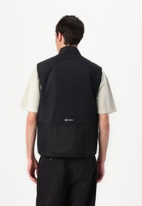 Svart ermeløs vest laget av lett materiale med en teksturert mesh-lomme og kragedetalj. Designet med en avslappet passform og enkel silhuett.