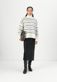 Übergroßer weißer Pullover mit schwarzen horizontalen Streifen, kombiniert mit einem schwarzen Midi-Rock und kniehohen schwarzen Stiefeln. Braune Handtasche in der Hand.