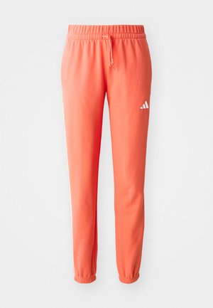 Korallfargede sweatshirtbukser med elastisk linning og snøring. Har hvit Adidas-logo og to sidestriper. Stoffet er mykt med en jevn tekstur.