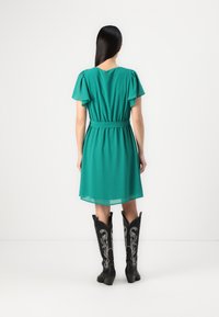 Robe verte à manches courtes avec une taille cintrée, un tissu fluide et un ourlet à hauteur du genou, associée à des bottes de cowboy noires avec des coutures blanches intricacies.