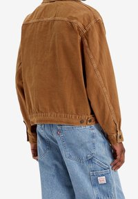 Osoba w brązowej kurtce i jasnoniebieskich dżinsach z łatą na kieszeni uda, stojąca tyłem.