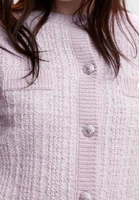 Světle růžový pletený cardigan s texturovaným vzorem, obsahující dvě přední kapsy a dekorativní knoflíky podél zapínání.