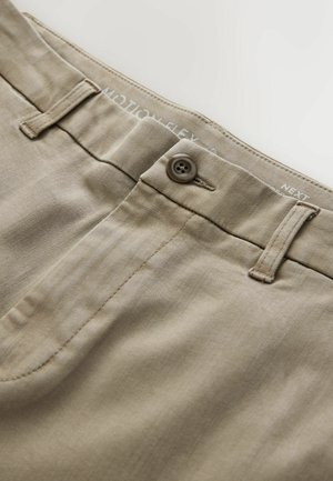 Bēškrāsas khaki bikses tuvplānā, kurā redzama poga, josta ar cilpām un daļa no priekšējā kabatas šuves.