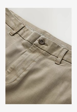 Bēškrāsas khaki bikses tuvplānā, kurā redzama poga, josta ar cilpām un daļa no priekšējā kabatas šuves.