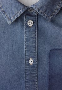 Kołnierzyk i listwa zaszewki koszuli dżinsowej z trzema widocznymi guzikami, jednym ozdobnym z przezroczystymi cyrkoniami oraz dwoma standardowymi guzikami.