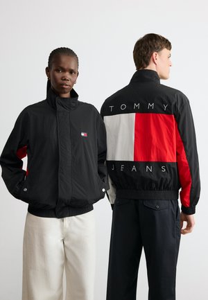 To modeller bærer sorte Tommy Jeans-jakker, den ene vender fremad med hvide bukser, den anden viser mærkenavnet på ryggen med sorte bukser.