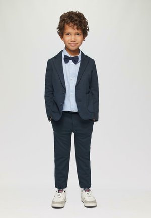 Marineblauw blazer en bijpassende broek over een lichtblauw gestreept shirt. Gekenmerkt door een zwarte vlinderdas en witte sneakers met grijze accenten.
