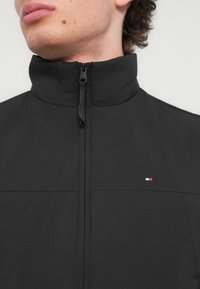 Μαύρο μπουφάν με φερμουάρ και ψηλό γιακά, με διακριτικό υφασμάτινο μοτίβο και μικρό τρίχρωμο λογότυπο στην αριστερή πλευρά κοντά στο φερμουάρ.