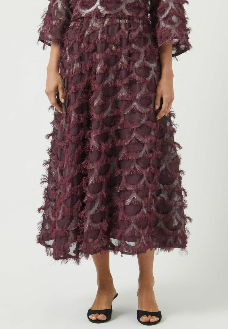 Mørkerød nederdel af let gennemsigtigt stof med lagdelte fjeragtige teksturer i et bølget mønster, med elastisk taljebånd og sorte sandaler.