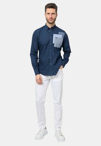 Camicia blu navy con tre toppe a motivi sul petto, abbinata a pantaloni bianchi e sneaker bianche. Tessuto morbido e traspirante.