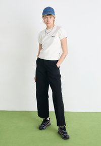 T-shirt bianco a coste con logo nero, abbinato a pantaloni neri a vita alta e gamba larga, e sneaker nere, completato con un cappellino in denim blu.