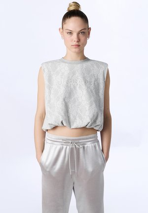 Jonge persoon met haar in een knot draagt een mouwloze grijze kanten top met de tekst "UPGRADE" en lichtgrijze broek met trekkoord, handen in de zakken.