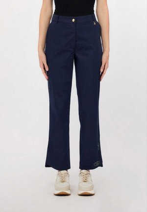 Donna che indossa pantaloni dritti blu navy con bottone dorato e ciondolo, abbinati a sneakers bianche, in piedi su sfondo bianco.