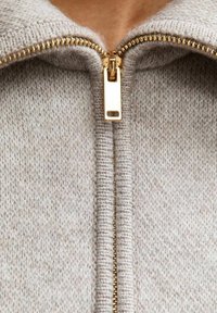 Suéter de color gris claro con cremallera, que presenta una cremallera dorada prominente y un tejido suave y texturizado, mostrando una vista en primer plano del cuello y la cremallera.