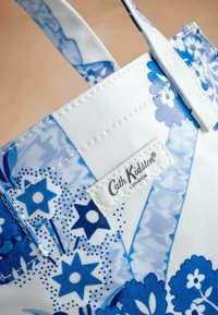 Baltā auduma soma ar zilu ziedu rakstu, kurai ir Cath Kidston etiķete. Materiāls izskatās teksturēts ar izšūtiem elementiem un zilām akcentiem.