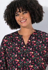 Blouse noire à motifs floraux avec décolleté en V, fermeture à boutons et motifs floraux multicolores vibrants sur un tissu lisse.