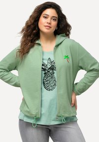 Haut à capuche zippé vert clair en tissu texturé, avec une petite broderie de palmier. En dessous, un t-shirt vert menthe avec un motif d'ananas.