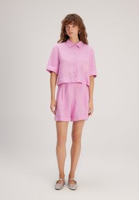 Camicetta rosa in lino con bottoni, tasca a toppa e dettagli ricamati, abbinata a pantaloncini rosa coordinati. Scarpe slip-on argentate.
