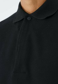 Polo shirt nera in tessuto strutturato, con colletto classico e chiusura a singolo bottone sul collo. Design semplice e funzionale.