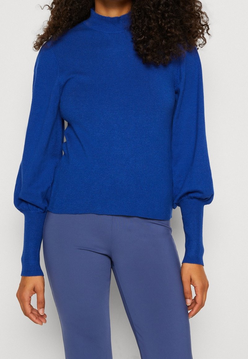 Blå stickad tröja med hög hals och puffärmar, textil med struktur, åtsittande vid handlederna, ihop med figurformande blå byxor.