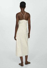 Robe longue beige clair à coupe droite, bretelles fines et un bouton décoratif dans le dos. Tissu texturé avec des motifs subtils.