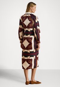 Kvinne iført en langermet midi-kjole med brunt, beige og svart geometrisk og kjede-mønster, kombinert med brune semskede flate sko.