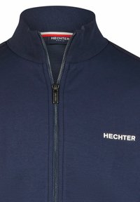 Marineblaue Zip-Jacke mit Stehkragen, aus glattem Stoff gefertigt. Besitzt einen Frontreißverschluss und das weiße "HECHTER"-Logo auf der Brust.