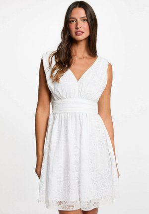 Femme portant une robe blanche en dentelle sans manches avec un col en V et une taille cintrée, debout devant un fond blanc uni.