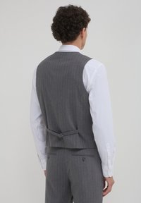 Homme avec des cheveux bouclés portant une chemise blanche et un gilet à rayures grises, vu de dos contre un fond uni.