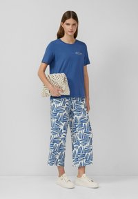 T-shirt bleu à manches courtes avec texte, associé à un pantalon large à motifs blancs et bleus. Le modèle tient une pochette tissée beige. Baskets blanches.