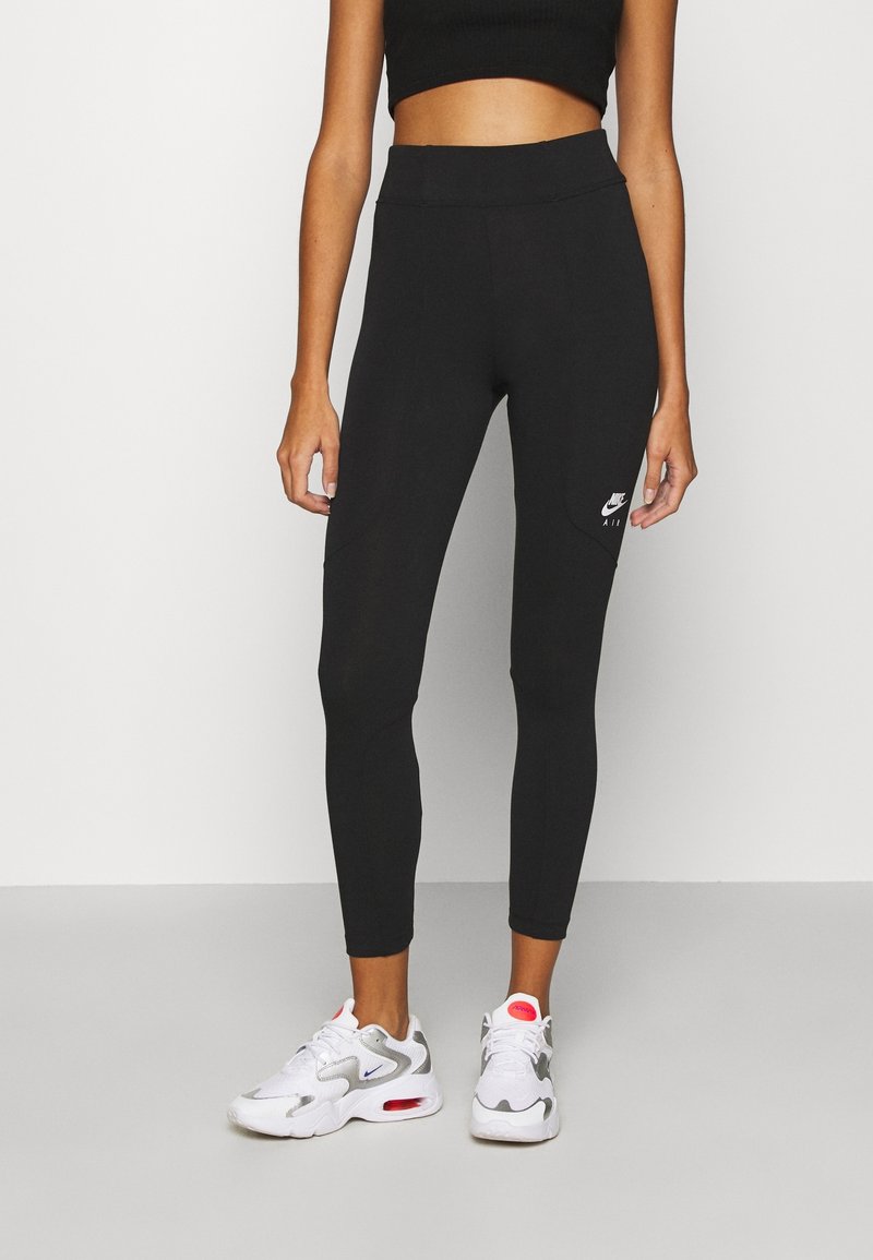 Kvinna i svarta Nike leggings och vita träningsskor med grå och röda detaljer, som står framför en ljus enkel bakgrund.