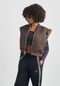 Καφέ μπουφάν τύπου bomber με γούνινες λεπτομέρειες, κλείσιμο με φερμουάρ και λογότυπο. Φορεμένο πάνω από μαύρο crop top και συνδυασμένο με μαύρο joggers με πλευρικές ρίγες.