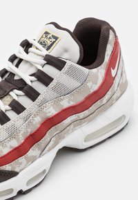 Sneaker mit beigem strukturiertem Obermaterial, rotem Wellenakzent, braunem Mesh-Zunge, weißem Nike Swoosh und sichtbarer Luftpolsterung.