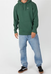 Sudadera verde con capucha y bolsillo delantero, combinada con jeans azul claro y zapatillas negras con detalles en blanco.
